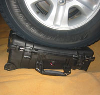 a close up of a black peli storm case in the sand with a car wheel on top of it